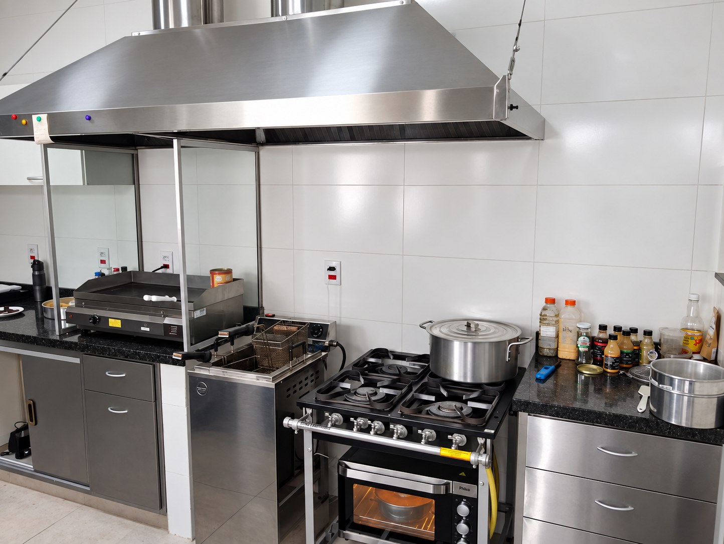 Cozinha industrial com coifa em aço inox, chapa, fritadeira e fogão sob bancadas de inox