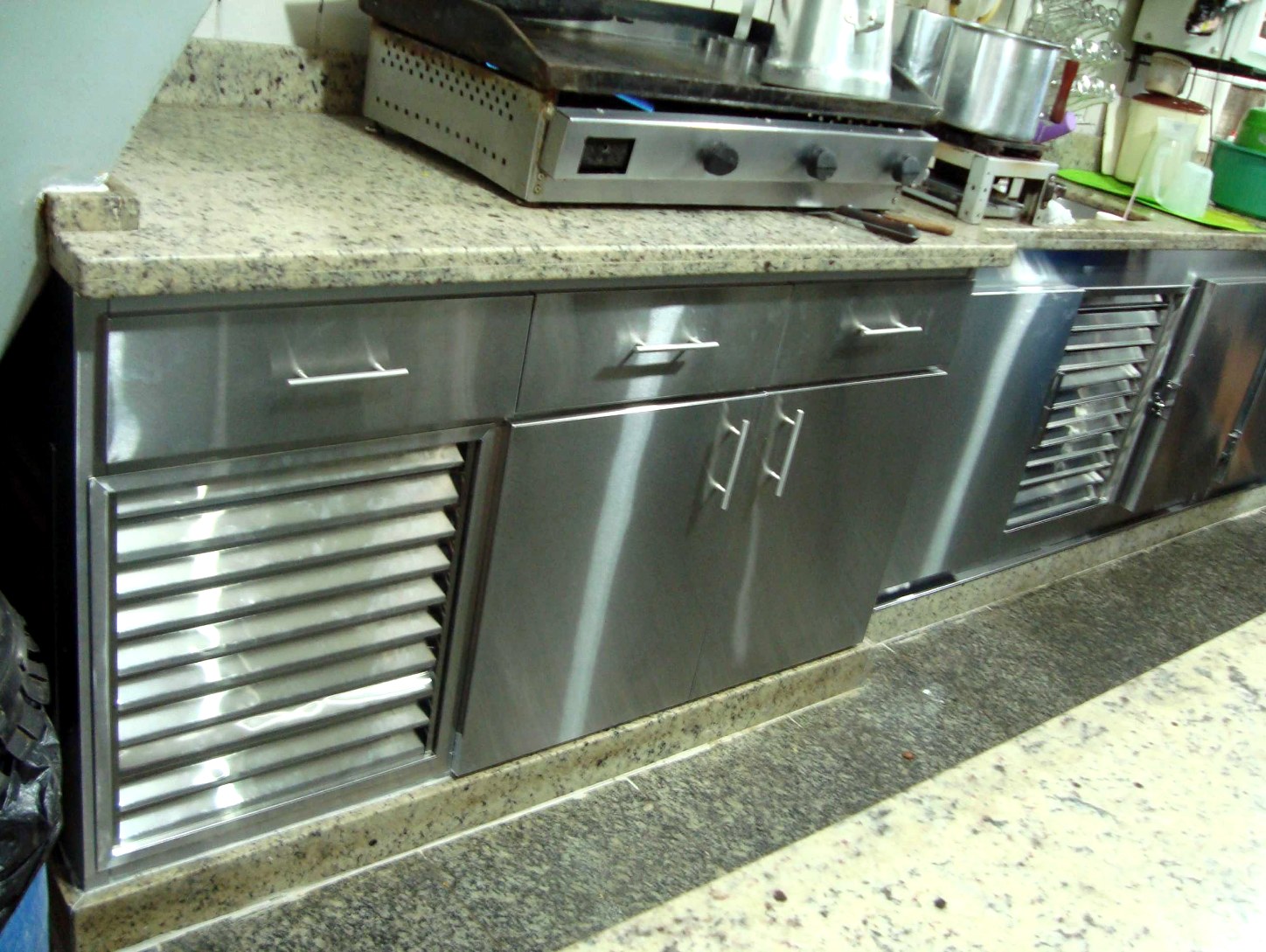 Fila de armários em aço inox com tampo de granito e linha de cocção, em cozinha comercial