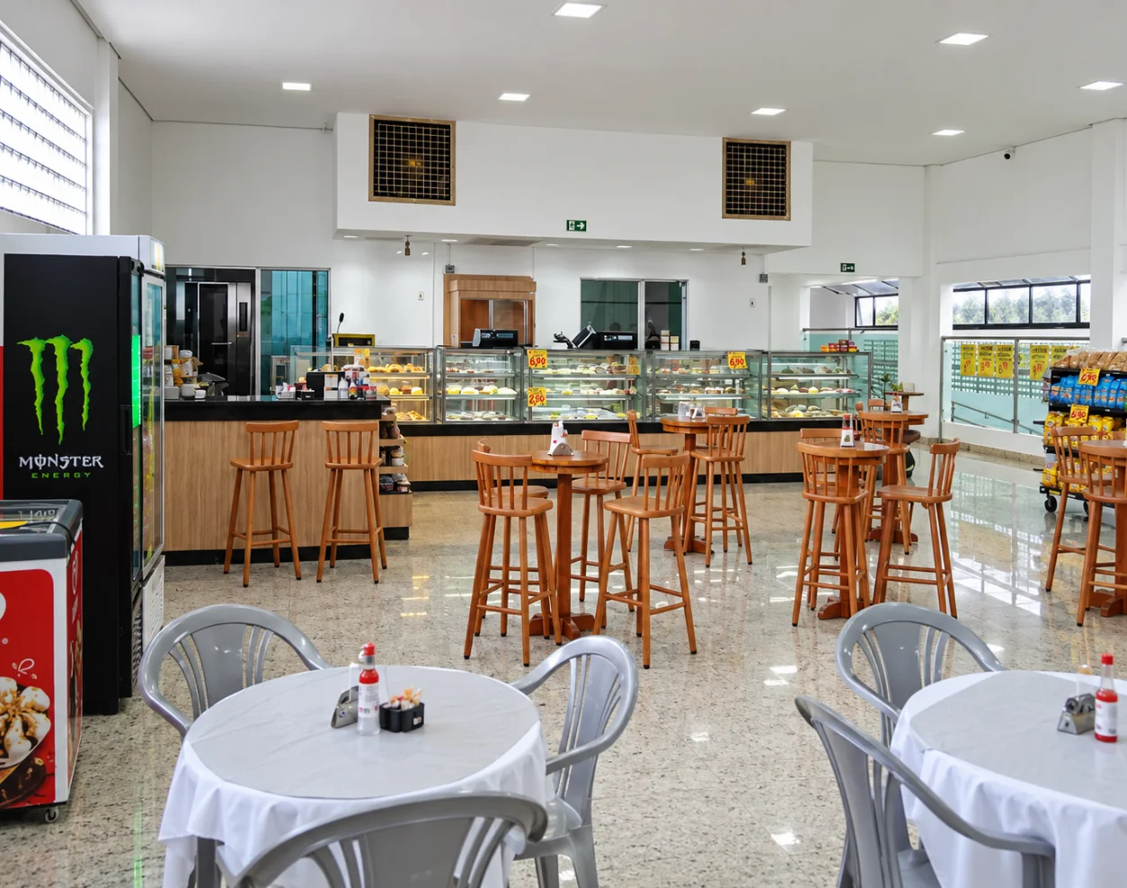 Salão amplo da padaria com mesas altas, balcão de atendimento e vitrines ao fundo