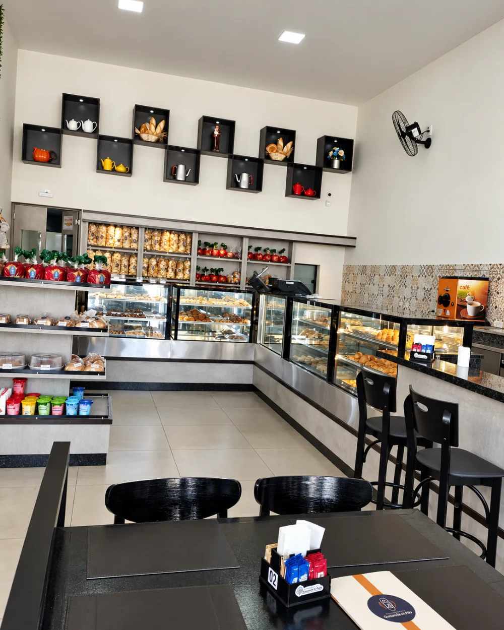 Salão com nichos iluminados na parede, balcão em L com vitrines e área de café
